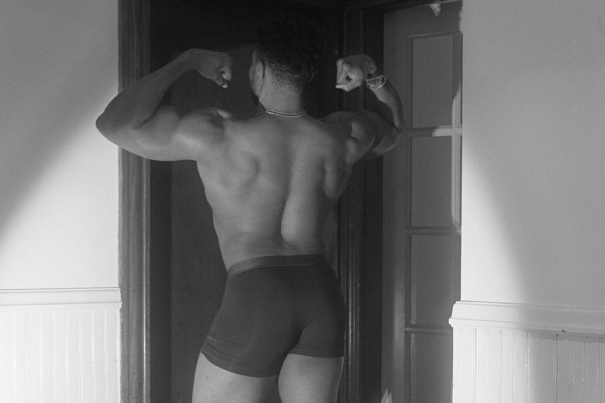 Photo by Megan Christiansen of a black man flexing in a doorway while wearing dark boxer shorts and evening light illuminatinghim and the wall